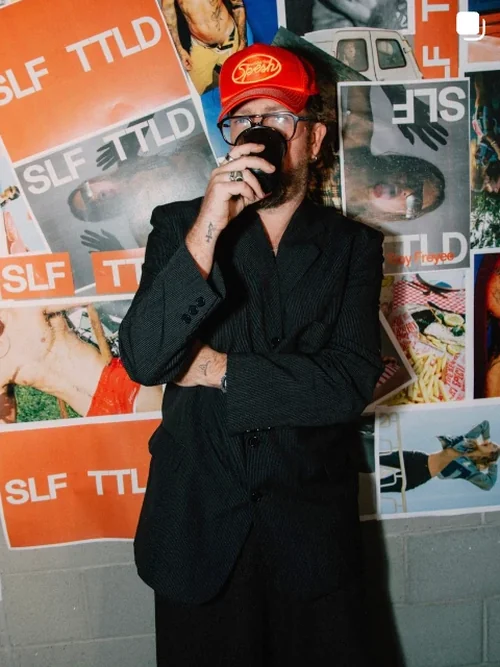 Made By Muse Instagram - Man in a red cap drinking from a mug in front of SLF TTLD posters